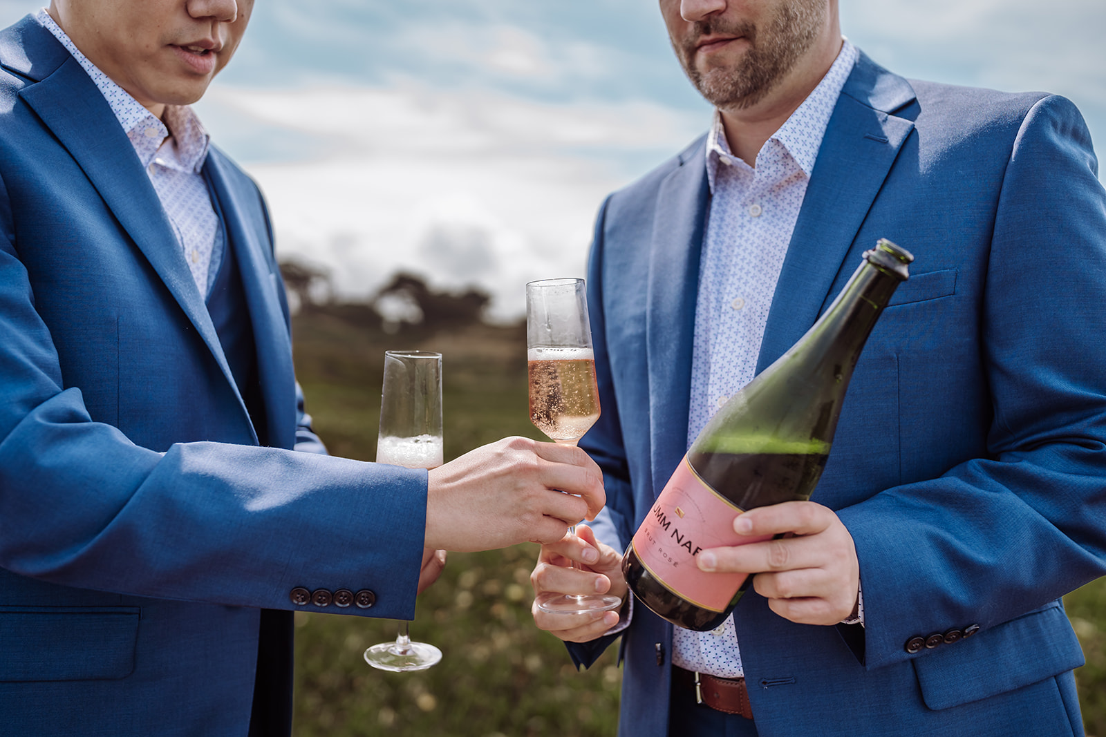 An inclusive and intimate wedding and elopement photographer, Xilo Photography, Kati Douglas who is based in Oakland, California in the bay area, took this picture of a same sex gay couple on their wedding day/elopement day as the two grooms pour themselves champagne to in champagne glasses while they celebrate their wedding day in their tuxes at Fort Funston, CA which was their intimate wedding venue. Ever wondered what is an intimate wedding or what would be considered a small wedding? This is a great example of both! 