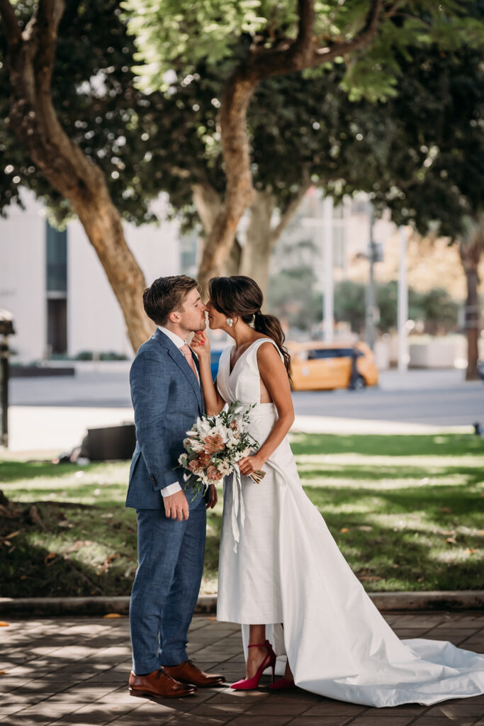 LA wedding that had a courthouse elopement as well as after party, reception, and vows with family. This picture was taken by San Francisco and Oakland Bay Area Photographer, Xilo Photography, Kati Douglas. She specializes in capturing gorgeous intimate and intentional weddings in and around the Bay Area. Pictured here is a groom in his classic blue tailored wedding tux, and the bridge in a gorgeous hi-low romantic and modern wedding dress. They are facing each other and leaning in for a kiss while she holds her gorgeous wedding bouquet. They are standing on a sidewalk near the LA County Courthouse after they eloped.