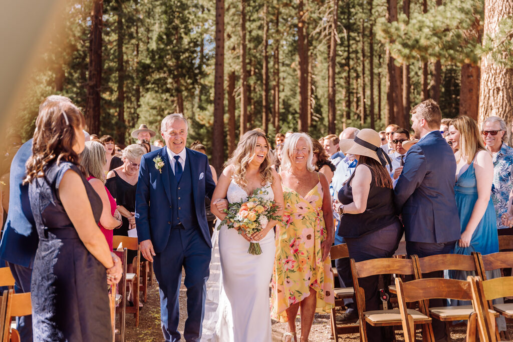 Lake Tahoe, California mountain, forest and beach wedding with the most beautiful views. This was considered a fairly intimate small wedding. The views did not disappoint and neither did the bride in her gorgeous romantic A-Line wedding dress. This image shows the wedding isle, the seating arrangements for the vow exchange and ceremony, guests, and the stunning bride, and her father and mother walking her down the isle to get married. She is holding her colorful bouquet in her hand. Photographed by Bay Area photographer, Kati Douglas with Xilo Photography based in Oakland, CA