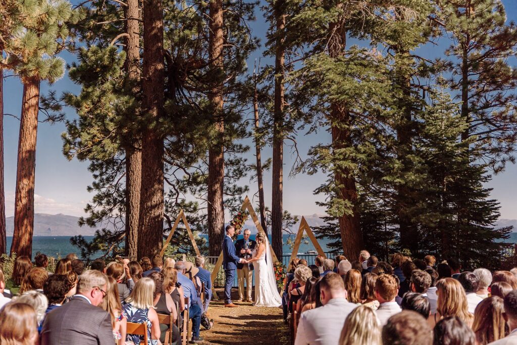 Lake Tahoe, California mountain, forest and beach wedding with the most beautiful views. This was considered a fairly intimate small wedding. The views did not disappoint and neither did the bride in her gorgeous romantic A-Line wedding dress. This image shows the wedding isle, the seating arrangements for the vow exchange and ceremony, guests, and the stunning and unique mountain arch decor. Photographed by Bay Area photographer, Kati Douglas with Xilo Photography based in Oakland, CA