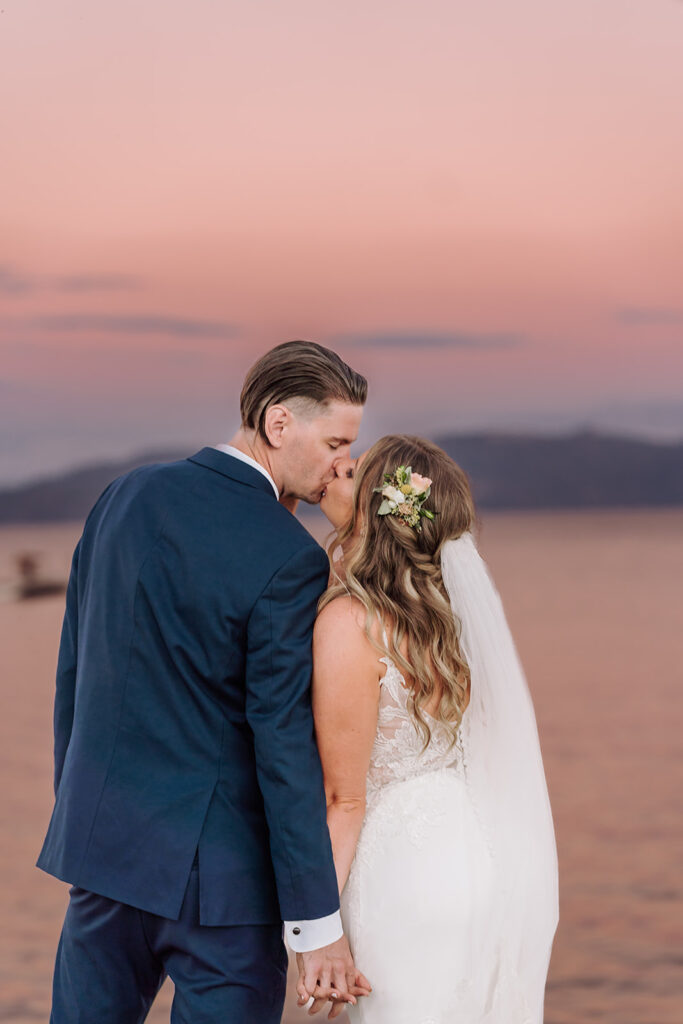 Pictured here is the bride and groom in their wedding tux and gorgeous gown, on the dock at Lake Tahoe, CA. They are kissing during the sunset and the skies are a mix of pinks, purple, and coral colors. This was taken in Lake Tahoe, California during a mountain, forest wedding and beach wedding. Photographed by Bay Area photographer, Kati Douglas with Xilo Photography based in Oakland, CA. 